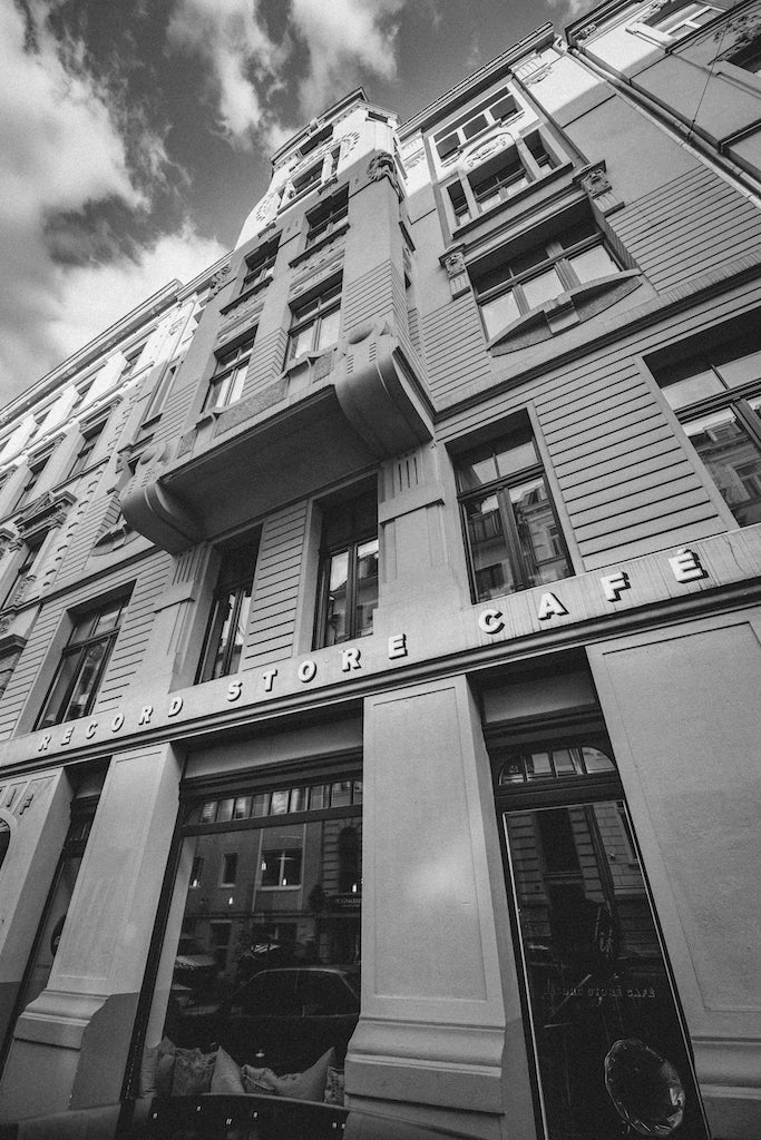 Schwarz-Weiß-Fotografie des as/if record store Cafés. Ein imposantes historisches Gebäude mit großen Fenstern und verzierter Fassade, die den Schriftzug 'Record Store Café' trägt. Die Architektur des Gebäudes verleiht der Szene einen klassischen und urbanen Charme, während der Himmel im Hintergrund teilweise von Wolken bedeckt ist.